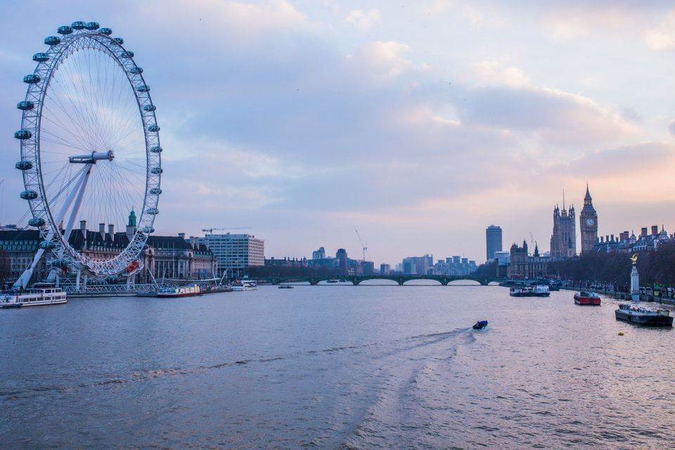 Una grande ruota panoramica e il paesaggio urbano lungo un ampio fiume con barche che navigano sotto un cielo crepuscolare.