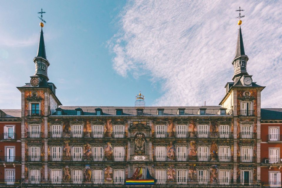 Facciata di edificio storico con elaborati affreschi murali, balconi multipli e torri dell'orologio a doppia guglia sotto un cielo azzurro.