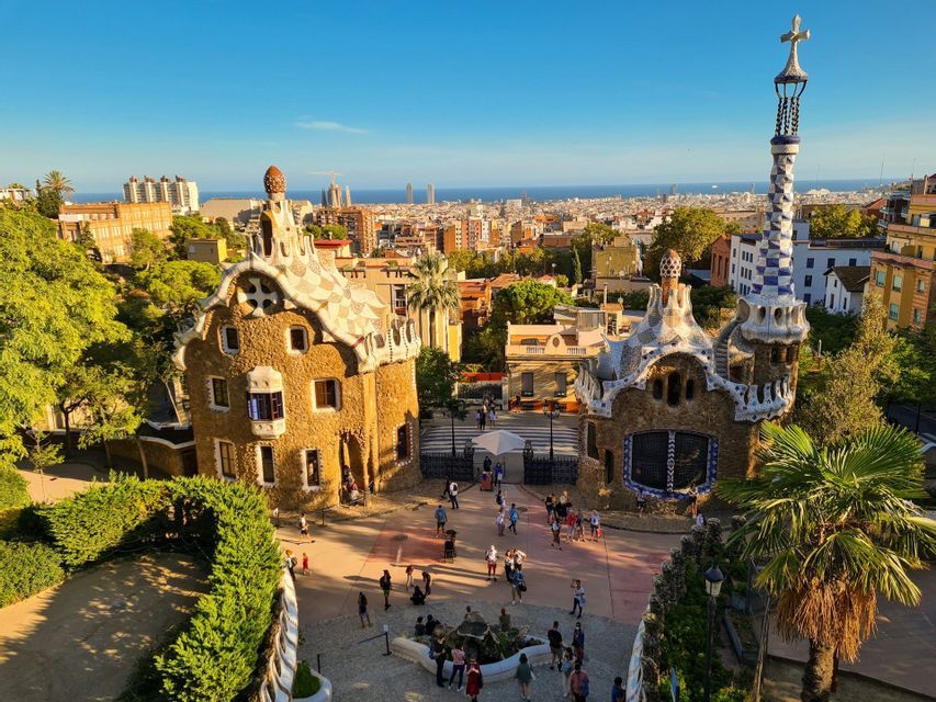 Visitantes caminando por ornamentados pabellones de piedra con agujas de mosaico con vistas a la ciudad y al mar bajo un cielo despejado.