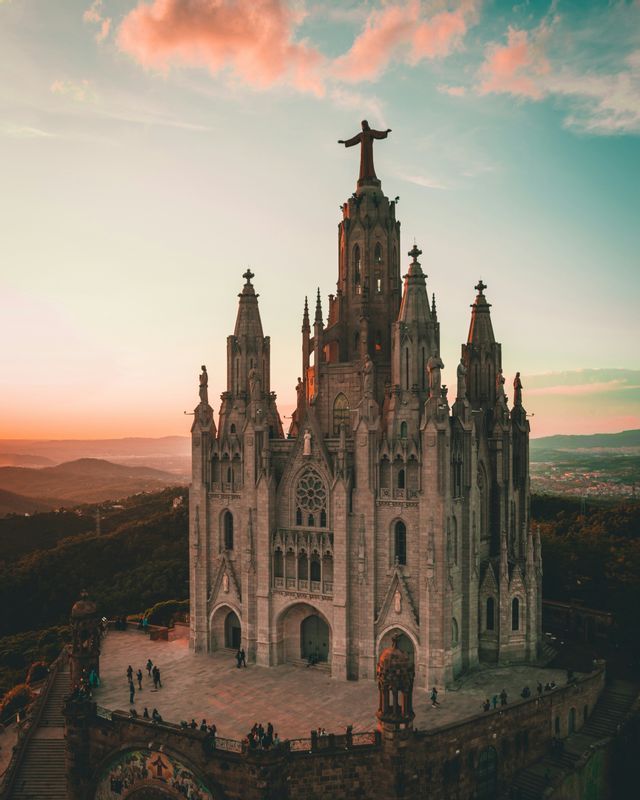 Una catedral de piedra con altas agujas y una estatua en la cima se alza sobre una colina contra un cielo al atardecer.