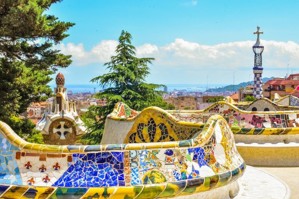 Bancos curvos de mosaico colorido dominan el extenso horizonte de la ciudad y el mar lejano bajo un cielo azul.