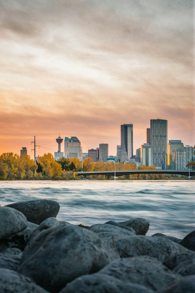 A city skyline rises behind a flowing river and a rocky bank under a cloudy sunset sky.