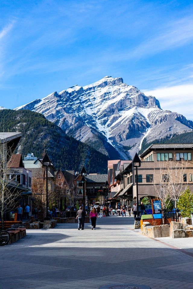 People walk down a paved pedestrian street in a town against the backdrop of a large snow-covered mountain peak.