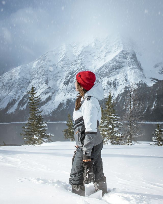 A person in winter clothing and a red beanie stands in deep snow overlooking a lake and snow-covered mountain.