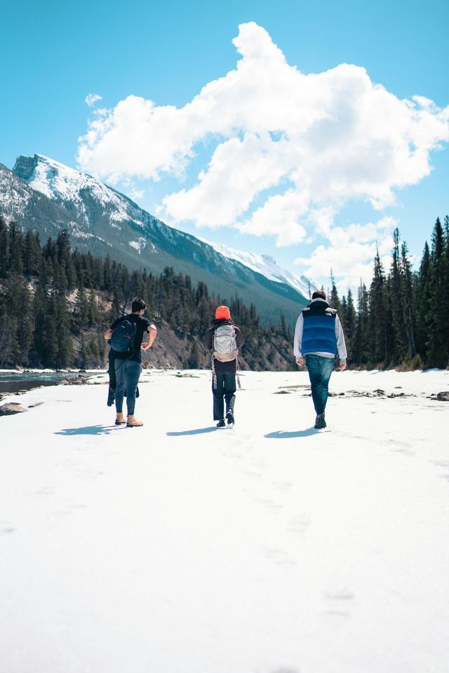 A WeRoad group trip of three people walking on snow along a river valley framed by pine trees and mountains.