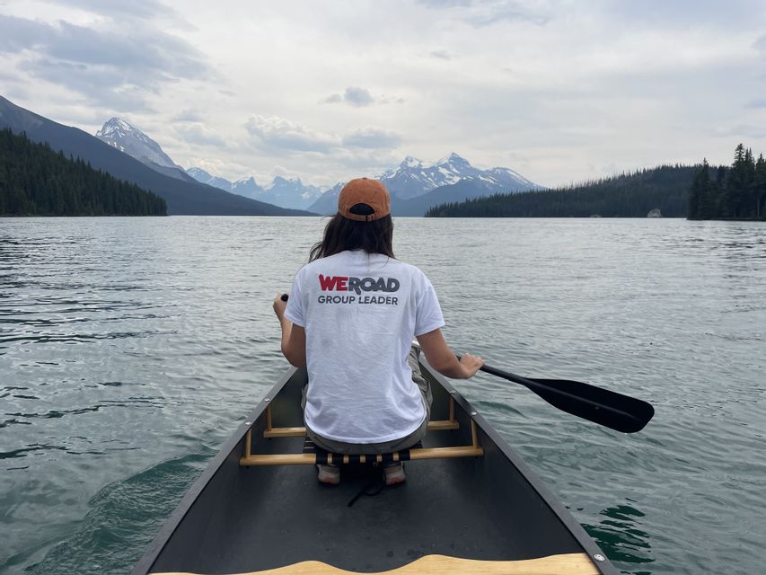 Un coordinatore di WeRoad pagaia in kanoa attraverso un ampio lago circondato da rive bosco-secche e cime montuose.