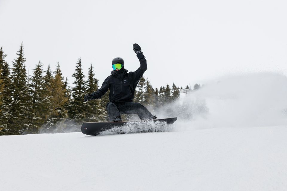 Un snowboarder vestido de negro descendiendo por una pendiente nevada, levantando nieve polvo fresca con pinos al fondo.