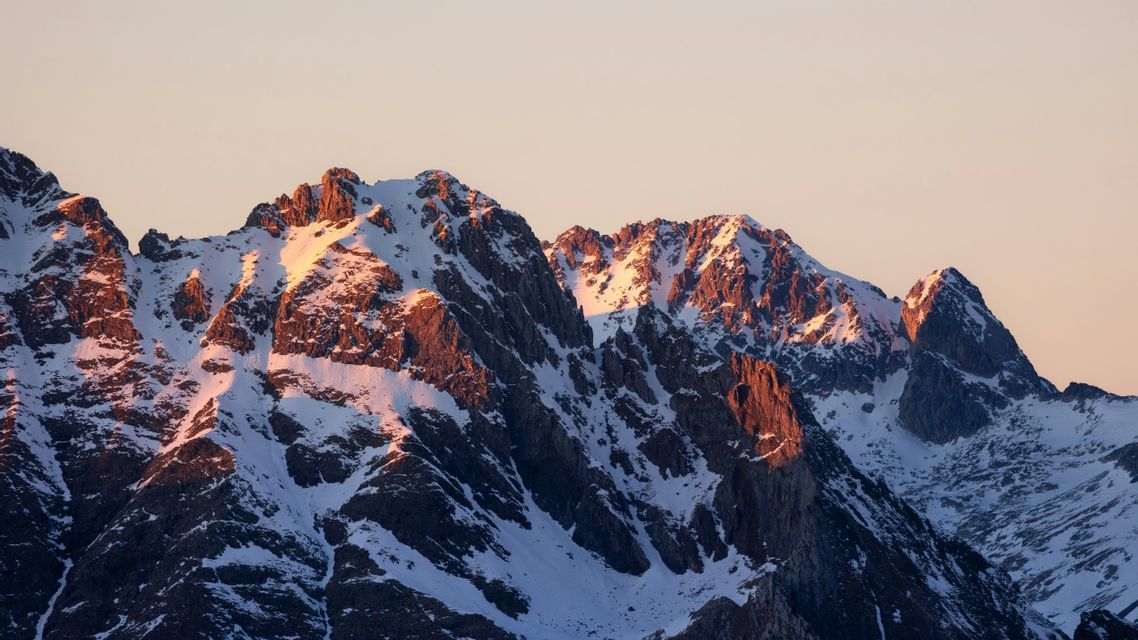 Picos de montañas rocosas cubiertas de nieve iluminados por una cálida luz solar dorada contra un cielo pálido.