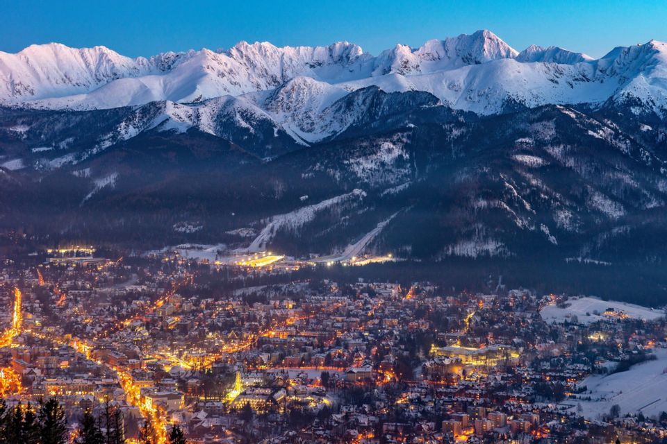 An illuminated town glows at dusk beneath towering snow-covered mountain peaks and forested slopes.
