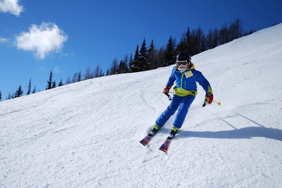 A skier wearing a helmet and blue ski suit skiing down a snowy mountain slope under a clear blue sky.