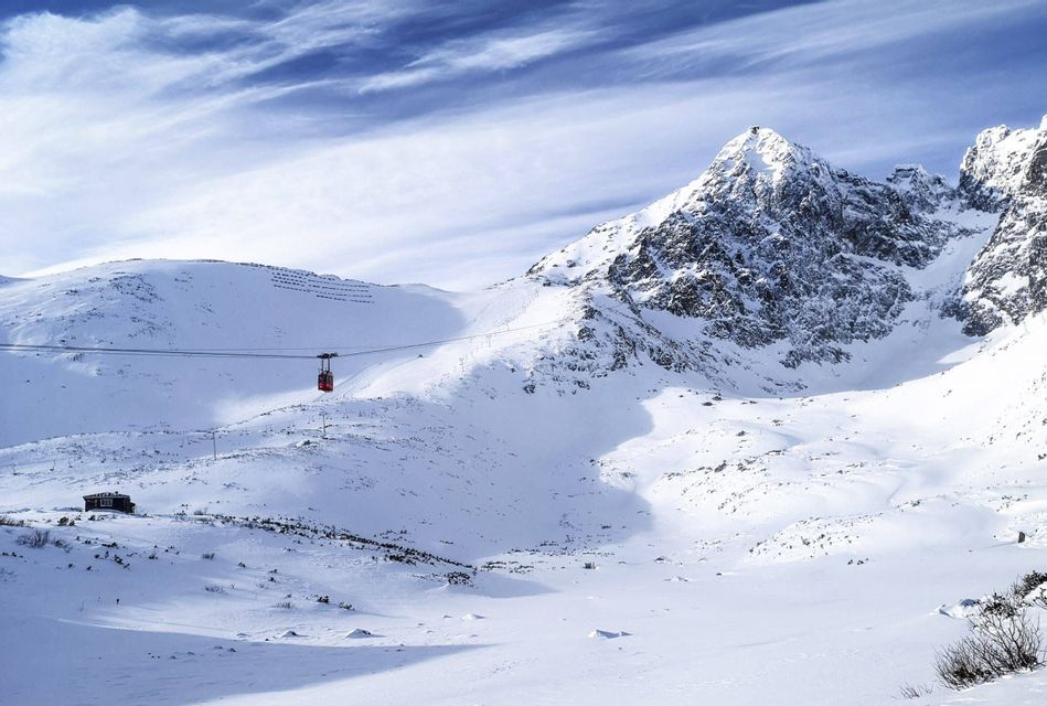 A red cable car travels across a snow-covered mountain valley under a partly cloudy blue sky.