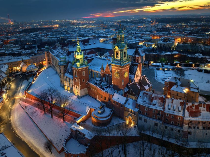 An illuminated historic brick castle with towers stands covered in snow overlooking a city at dusk.