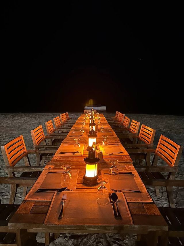 Un lungo tavolo da pranzo in legno apparecchiato con lanterne luminose, bicchieri e posate su una spiaggia buia di notte.