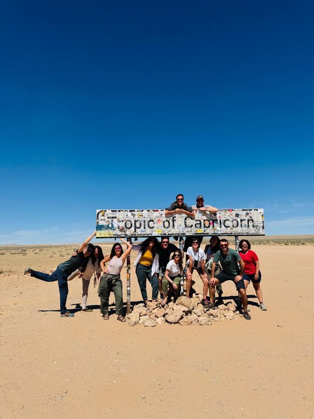 Un viaggio di gruppo WeRoad in posa vicino al cartello del Tropico del Capricorno in un deserto arido sotto un cielo azzurro