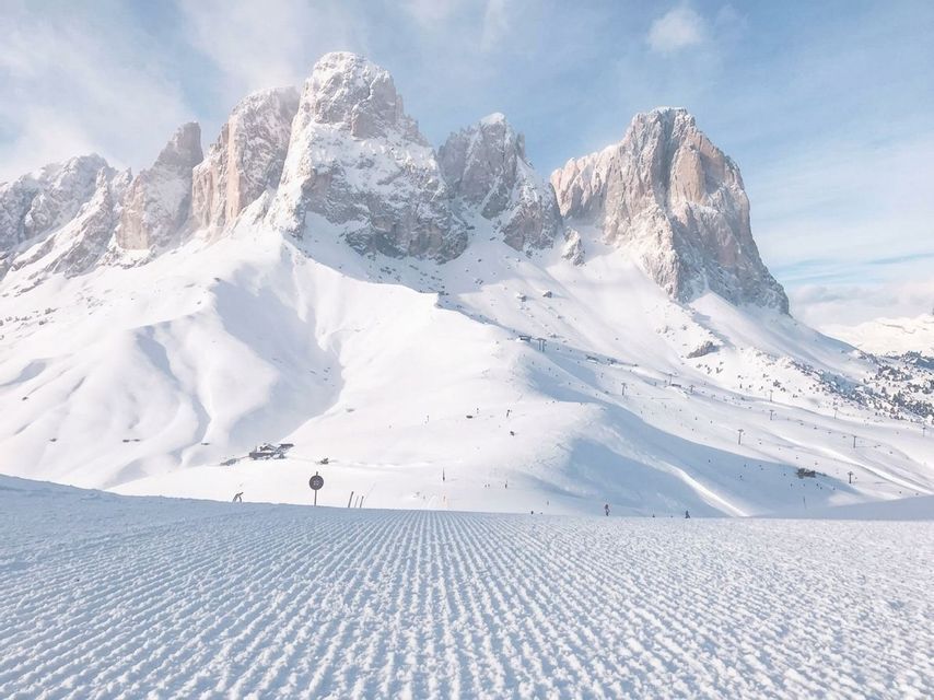 Neve battuta su una pista da sci che conduce verso imponenti cime montuose rocciose innevate sotto un cielo limpido.