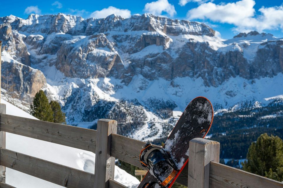 Uno snowboard è appoggiato a una ringhiera di legno che si affaccia su montagne aspre e innevate sotto un cielo azzurro.