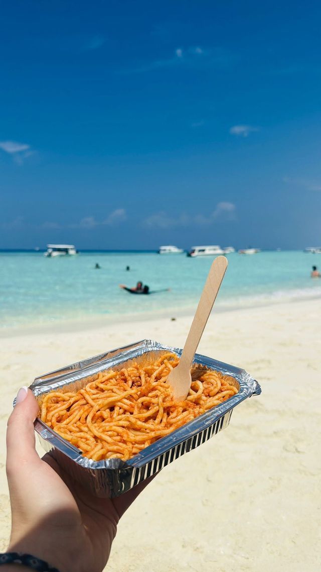 Una mano che tiene un contenitore di alluminio con spaghetti su una spiaggia di sabbia bianca e soleggiata con acqua turchese sullo sfondo