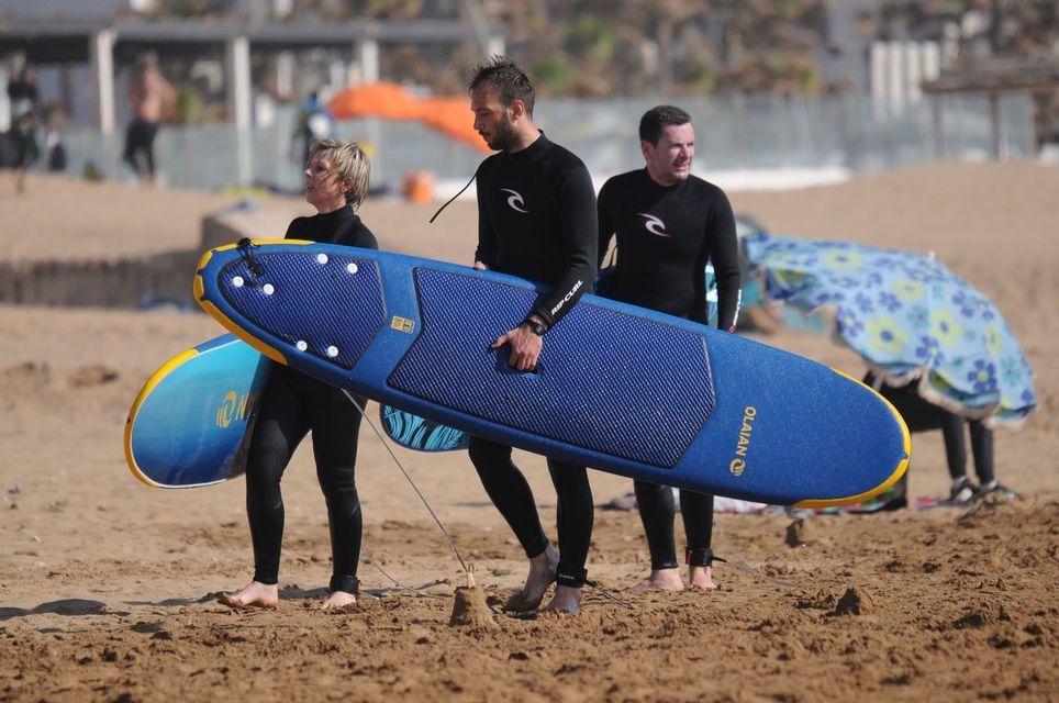 Un viaje en grupo de WeRoad con tres personas en trajes de neopreno llevando tablas de surf y caminando por una playa de arena.