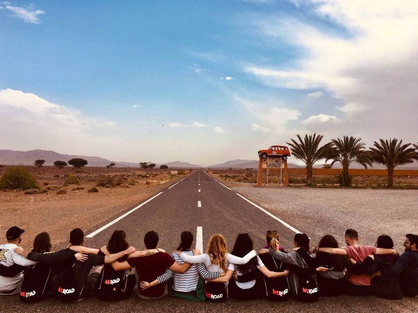 A WeRoad group trip seen from behind, sitting with arms around each other on an empty road in a desert landscape.