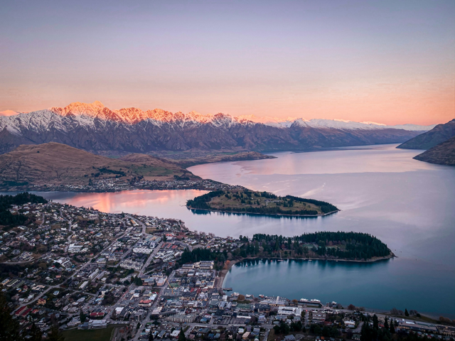 Vista aerea di una cittadina lacustre che confina con l'acqua e penisole boscose con montagne innevate che risplendono all'alba