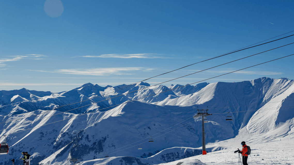Sciatori su una cresta di montagna innevata con seggiovie e cavi di telecabina contro cime innevate sotto un cielo azzurro.