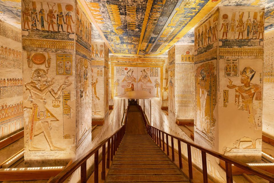 Una rampa di legno scende attraverso un'antica tomba fiancheggiata da colonne dipinte, geroglifici e colorati murales sul soffitto.