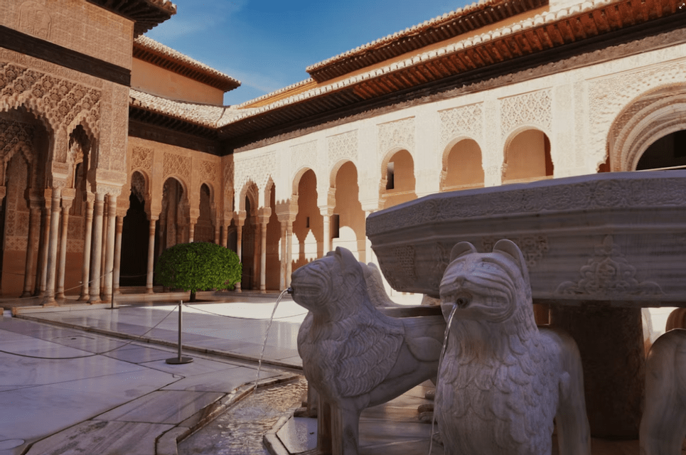 Una fuente de mármol con estatuas de leones arrojando agua en un patio soleado rodeado de intrincadas columnatas arqueadas.