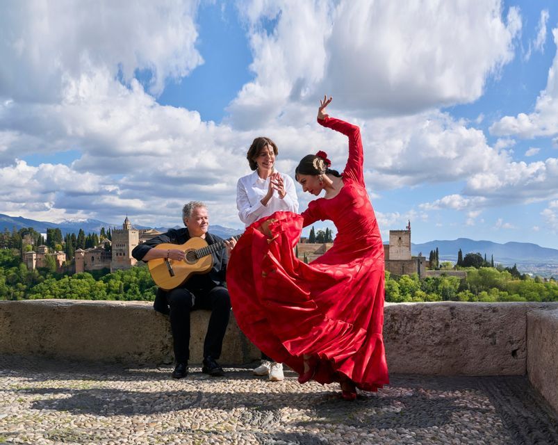 Una mujer con vestido rojo baila flamenco junto a un guitarrista y una mujer tocando las palmas al aire libre con vistas a una fortaleza histórica.