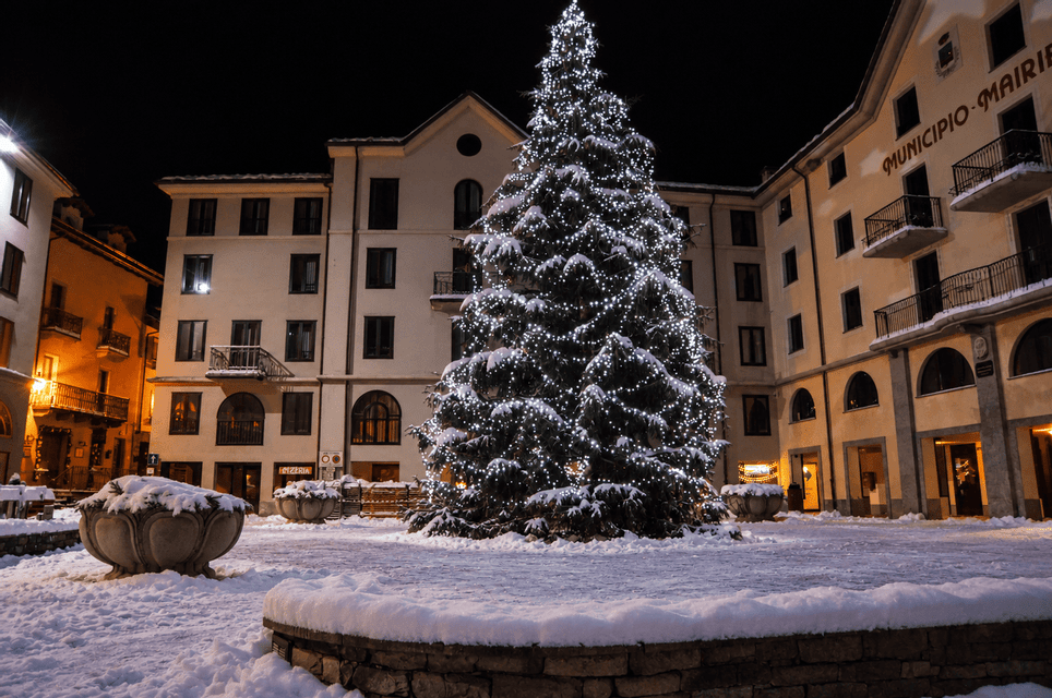Un grande albero di Natale illuminato si erge in una piazza di paese innevata di notte, circondato da edifici.