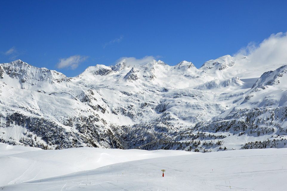 Cime montuose innevate si ergono dietro a una vasta pista da sci bianca sotto un cielo azzurro e limpido.