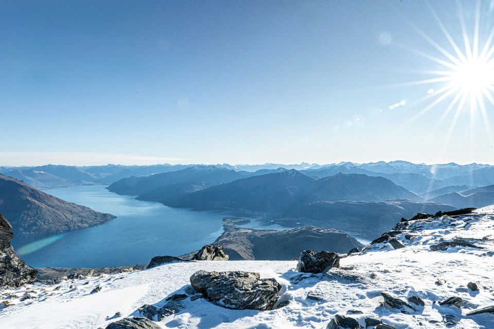Cima di montagna innevata che domina un grande lago serpeggiante e catene montuose lontane sotto un cielo luminoso e soleggiato.