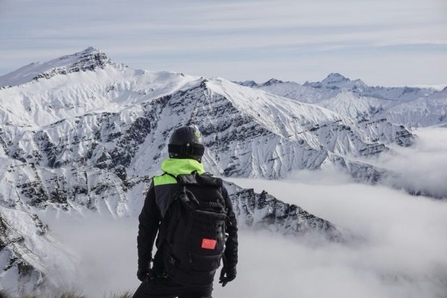 Una persona con uno zaino e un casco in cima a una montagna che ammira le cime innevate sopra un mare di nuvole