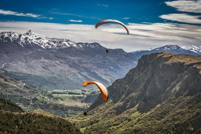 Due parapendii che sorvolano una valle verde circondata da montagne aspre con cime innevate sotto un cielo azzurro.