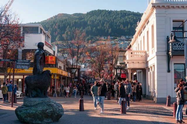 Persone che camminano lungo una strada pedonale fiancheggiata da negozi e da una statua, con una collina boscosa sullo sfondo.