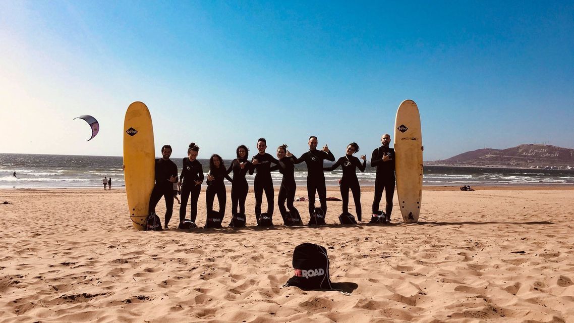 Viaje en grupo de WeRoad con trajes de neopreno, posando con tablas de surf en una playa de arena, con el mar y un cielo azul de fondo.
