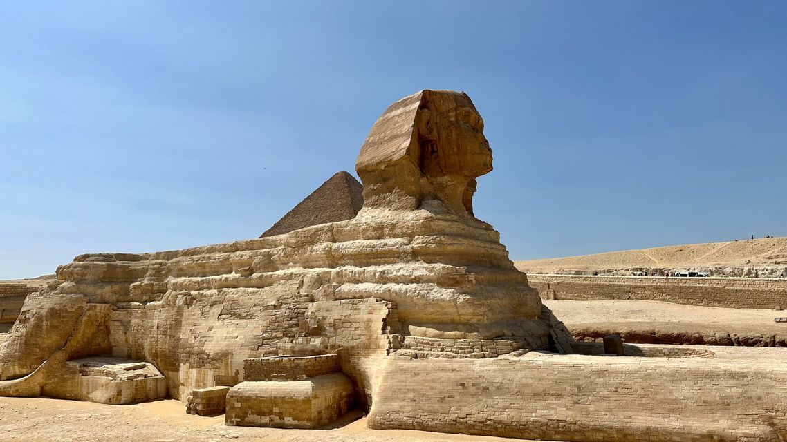 Seitenprofil eines massiven Steinsphingenmonuments in einer Wüstenlandschaft mit einer Pyramide im Hintergrund