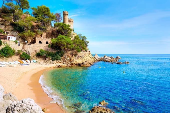 Un castello di pietra sorge su una scogliera rocciosa e alberata che domina una caletta sabbiosa con acqua di mare turchese sotto un cielo limpido