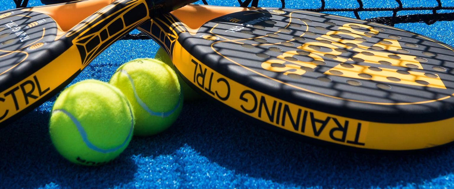 Due racchette da padel nere e gialle e palline verde brillante riposano su un campo in erba sintetica blu sotto l'ombra della rete