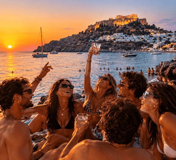 Un viaggio di gruppo WeRoad con drink su una spiaggia rocciosa al tramonto, con barche e un borgo collinare costiero sullo sfondo.