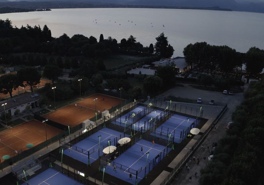 Illuminated blue padel courts and clay tennis courts beside a large lake at dusk, viewed from above.