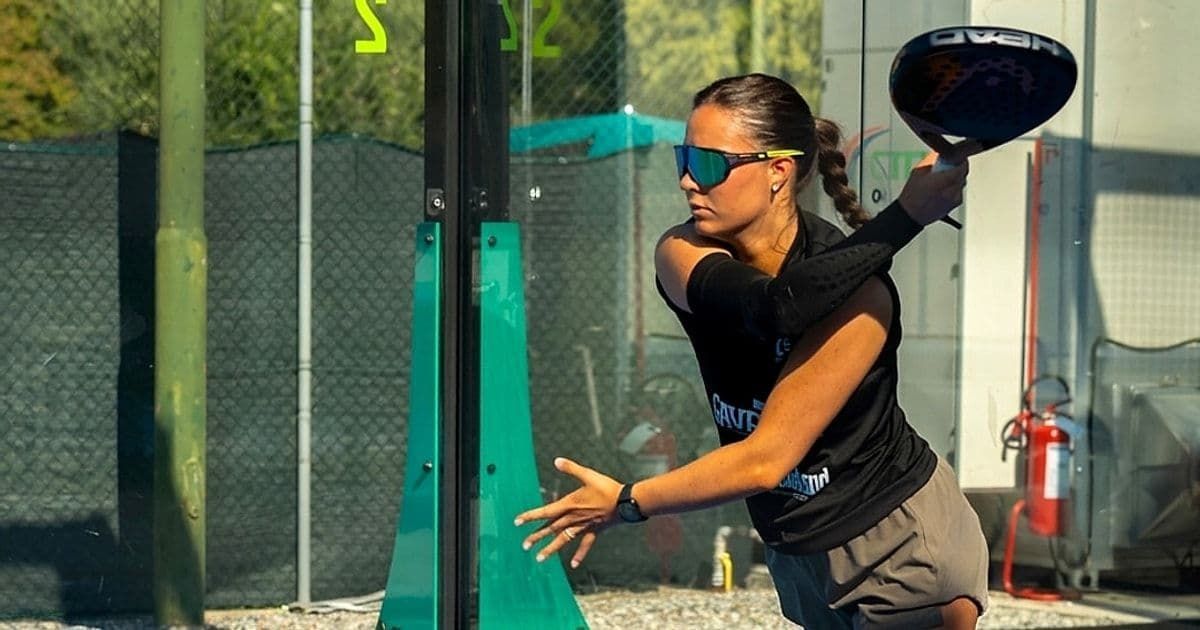 A woman wearing sunglasses prepares to hit a padel ball with her racket on an outdoor court.