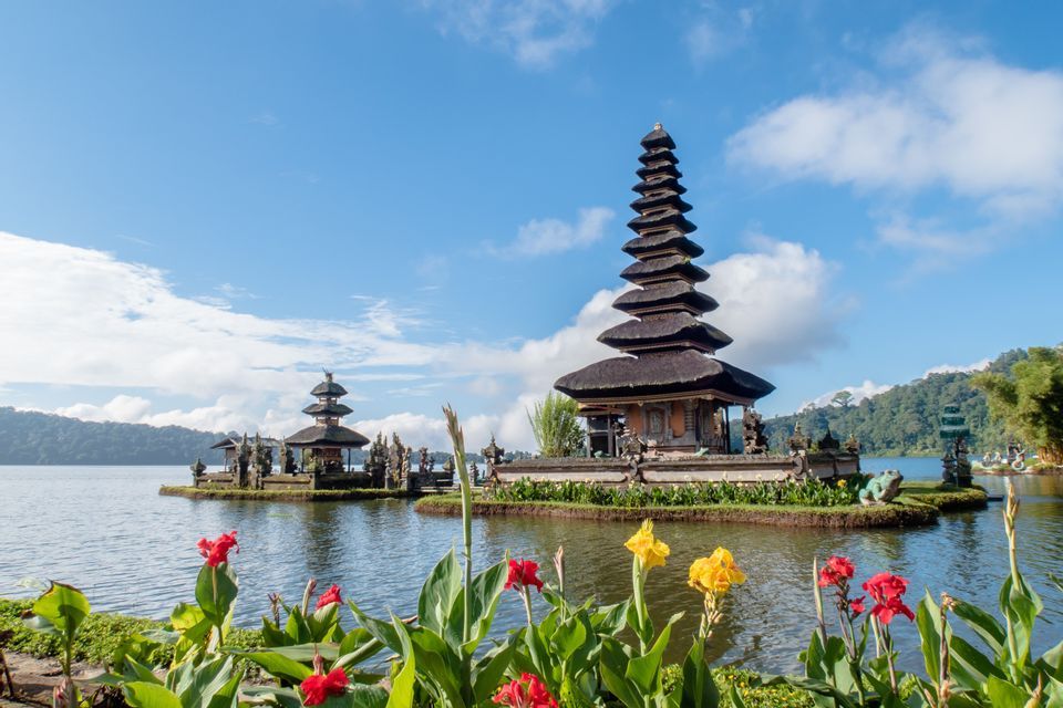 Two traditional temples with tiered roofs sit on a lake, with red and yellow flowers in the foreground under a partly cloudy sky.