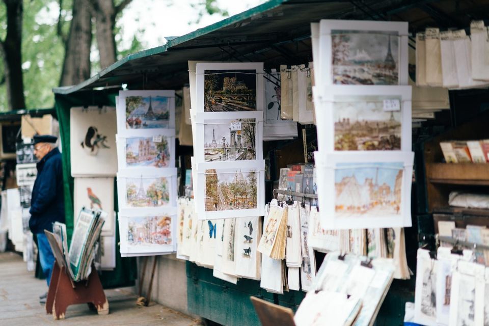 An outdoor stall displays numerous paintings and prints for sale, with a man in a beret standing to the side.
