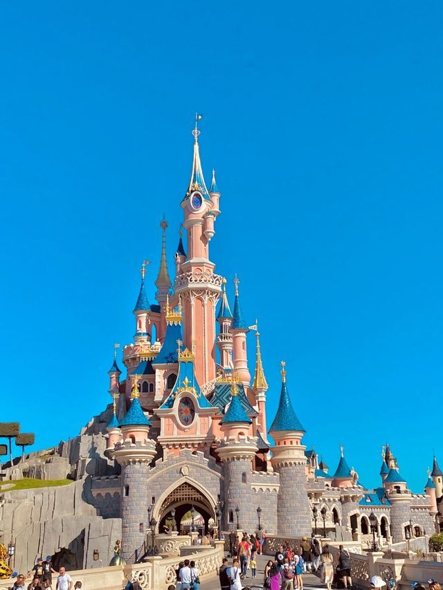 Un castello da fiaba rosa con torri dal tetto blu si erge imponente sotto un cielo azzurro limpido, con folle di persone alla sua base.