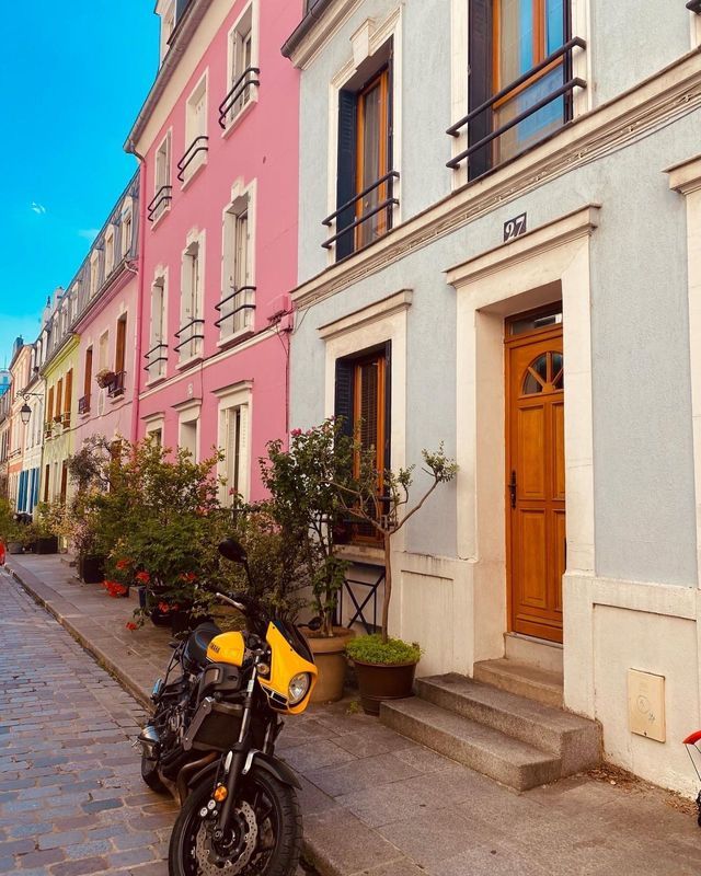 A yellow motorcycle is parked on a cobblestone street lined with colorful pink and blue apartment buildings.