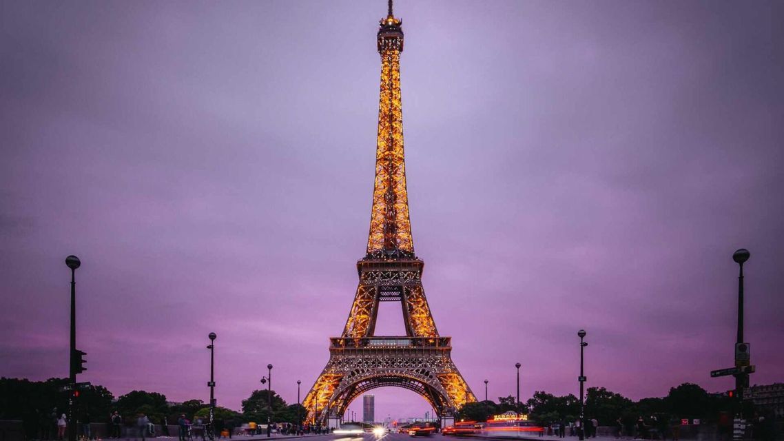 La Torre Eiffel, iluminada con luces doradas, se ve desde una calle con estelas de luces de tráfico bajo un cielo púrpura al anochecer.