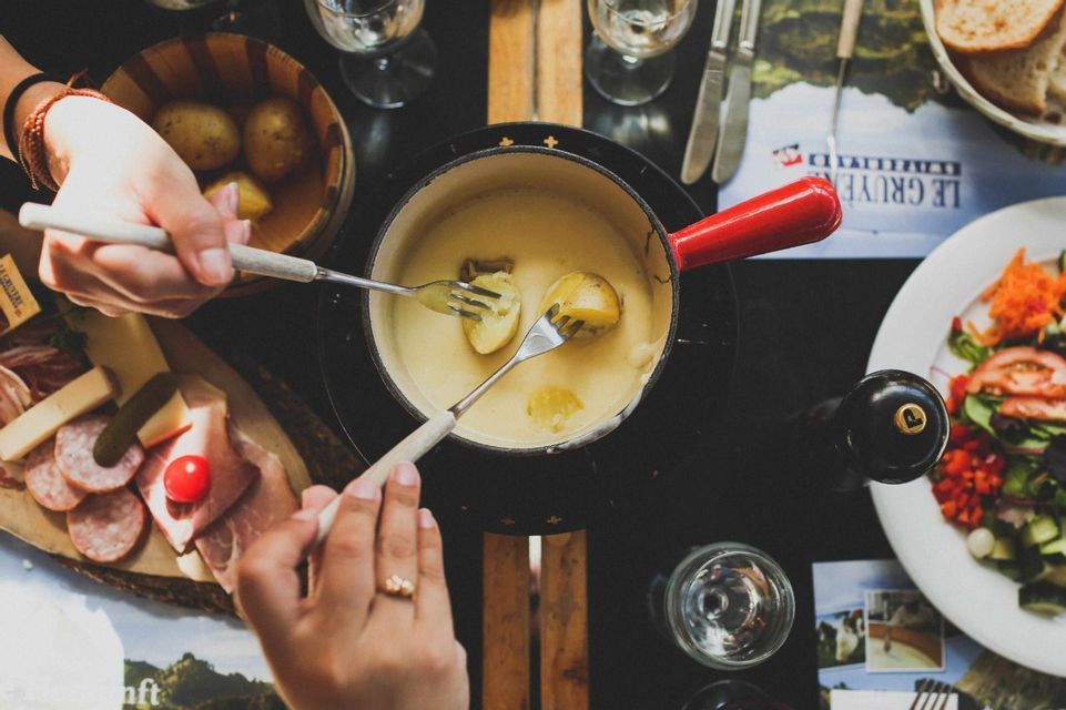 An overhead shot of two people dipping potatoes on forks into a shared pot of cheese fondue, with charcuterie on the side.