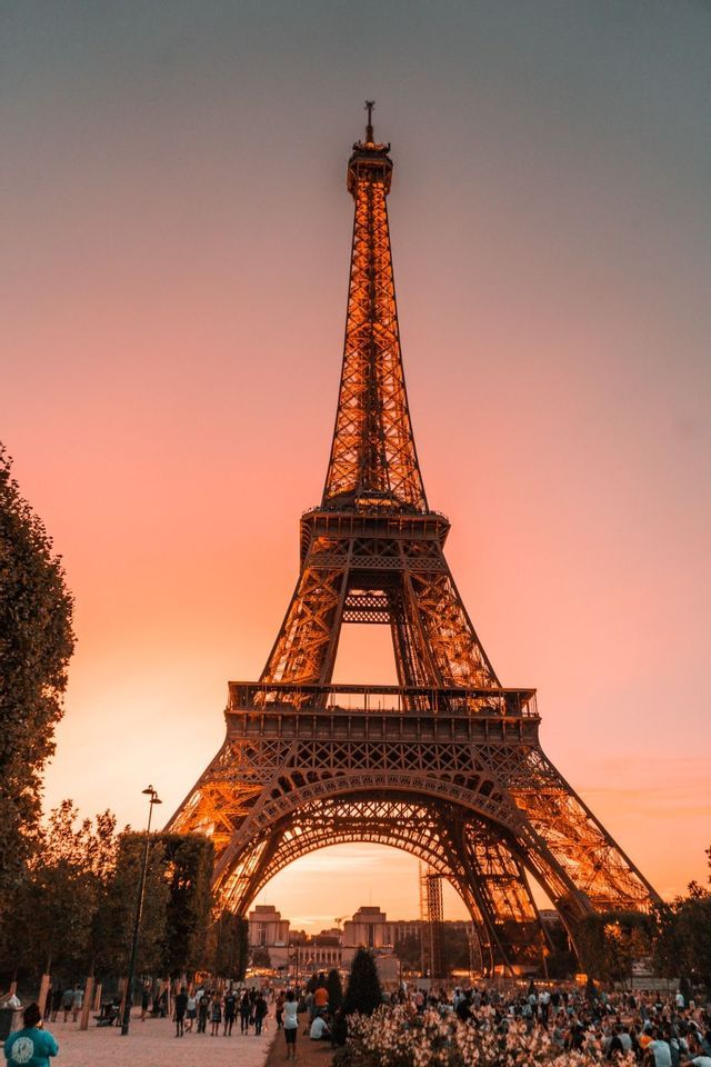 The Eiffel Tower seen from below against a pink and orange sunset sky, with crowds of people in the park at its base.