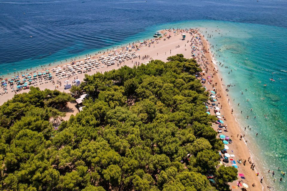 Vista aerea di una lingua di spiaggia triangolare fiancheggiata da pini e bagnanti circondata da acque limpide e blu.