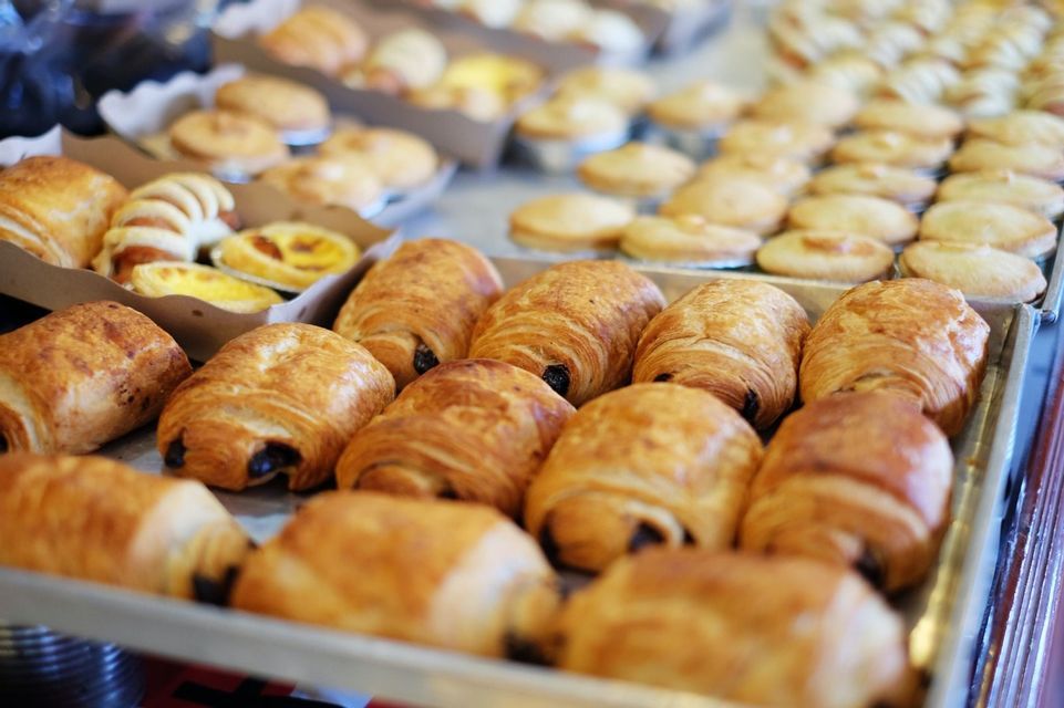 Trays of freshly baked pastries, with golden-brown chocolate croissants in the foreground of a bakery display.